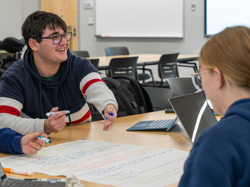two students talking and writing with markers in a classroom