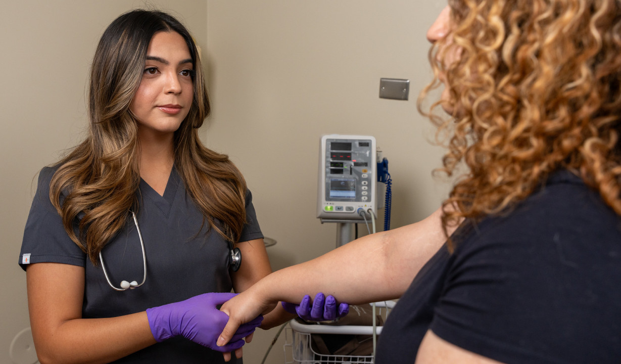 Student in a lab coat taking the pulse of another student in a clinical setting.